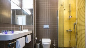 Un baño con lavamanos, espejo y retrete blancos, una ducha con azulejos amarillos y un cubo de basura.