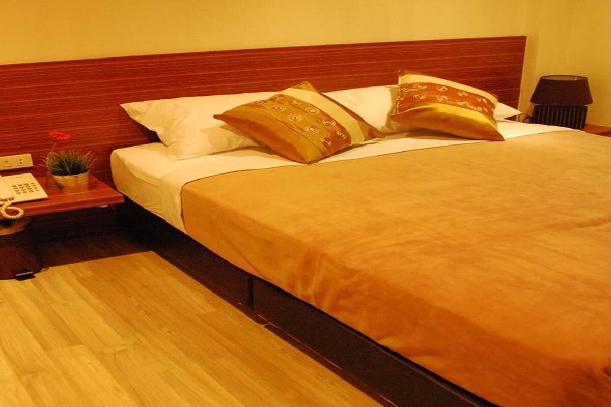 A hotel room with a wooden headboard, a bed with a beige bedspread and two patterned pillows, a nightstand with a telephone and a small plant, and a lamp on the right side.