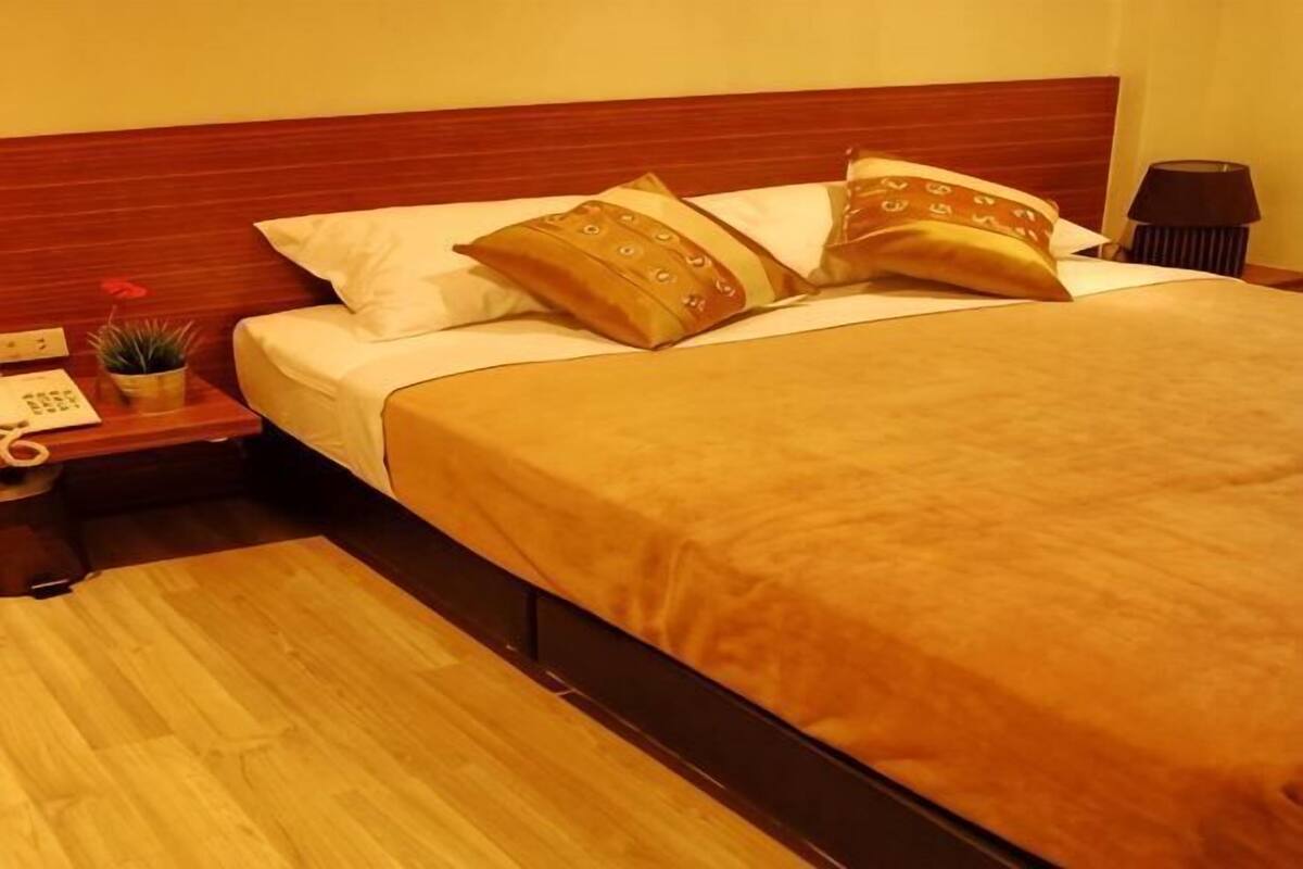 A hotel room with a wooden headboard, a bed with a brown bedspread, two decorative pillows, a nightstand with a phone and a small plant, and a lamp on the side table.