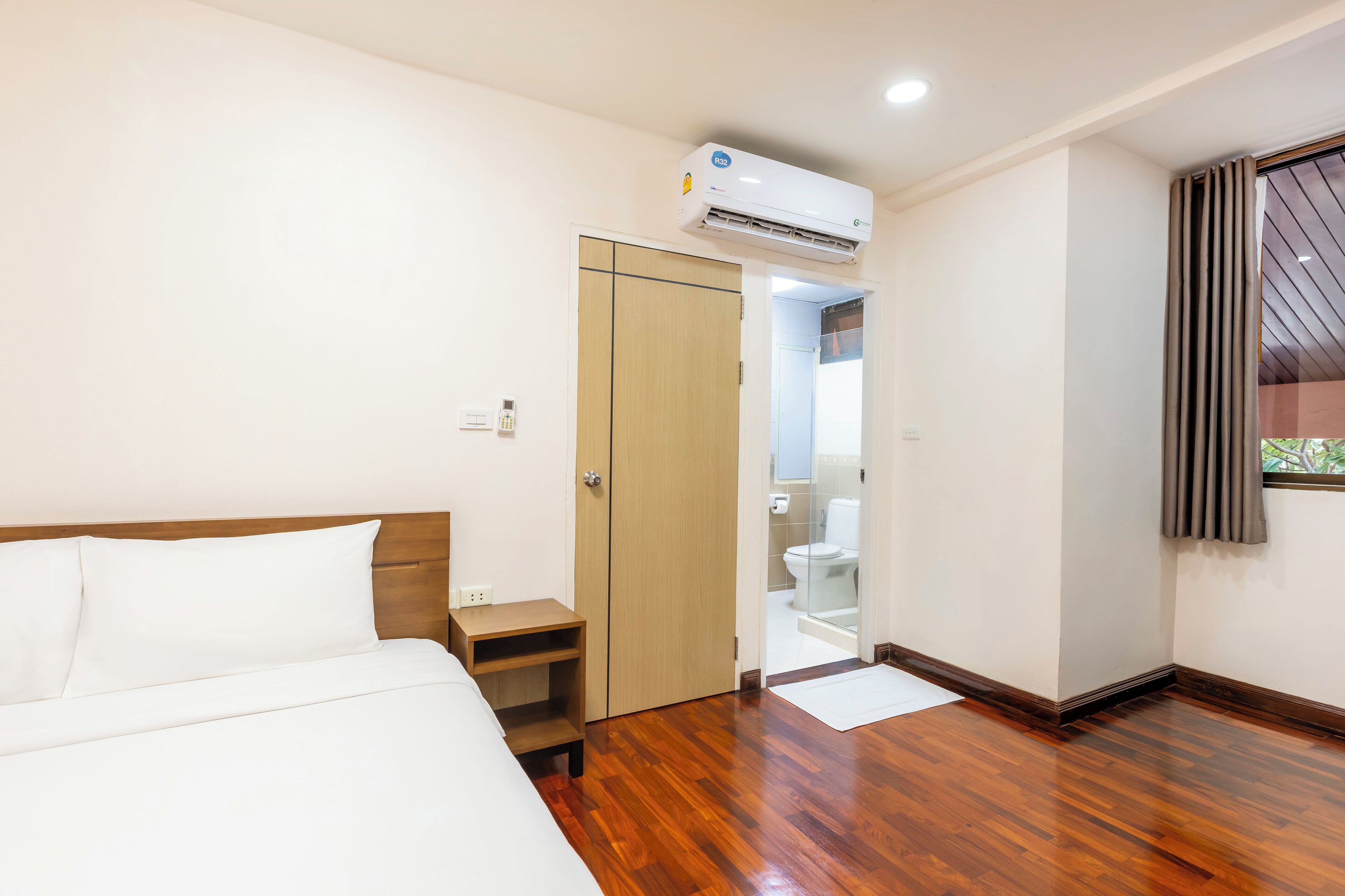 Ein Hotelzimmer mit Holzboden, einem Bett mit weißer Bettwäsche, einem Kleiderschrank, einer Klimaanlage und einem durch eine geöffnete Tür einsehbaren Badezimmer.