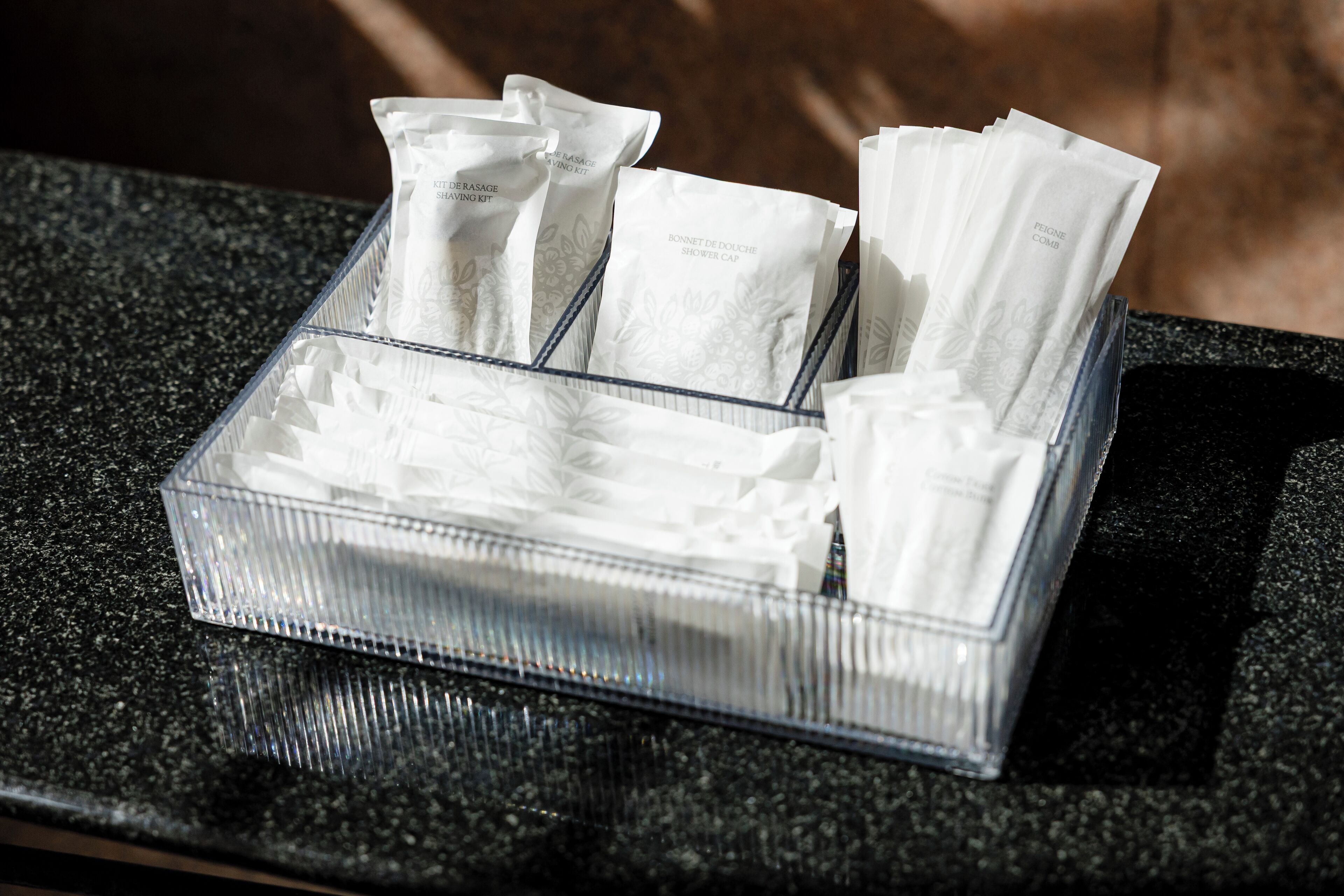 Un plateau en plastique transparent contient plusieurs sachets blancs étiquetés « BONNET DE DOUCHE ». Le plateau est posé sur un comptoir en granit.
