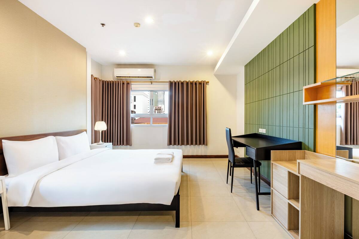 A hotel room with a bed, desk, chair, and a view of the city through the window.