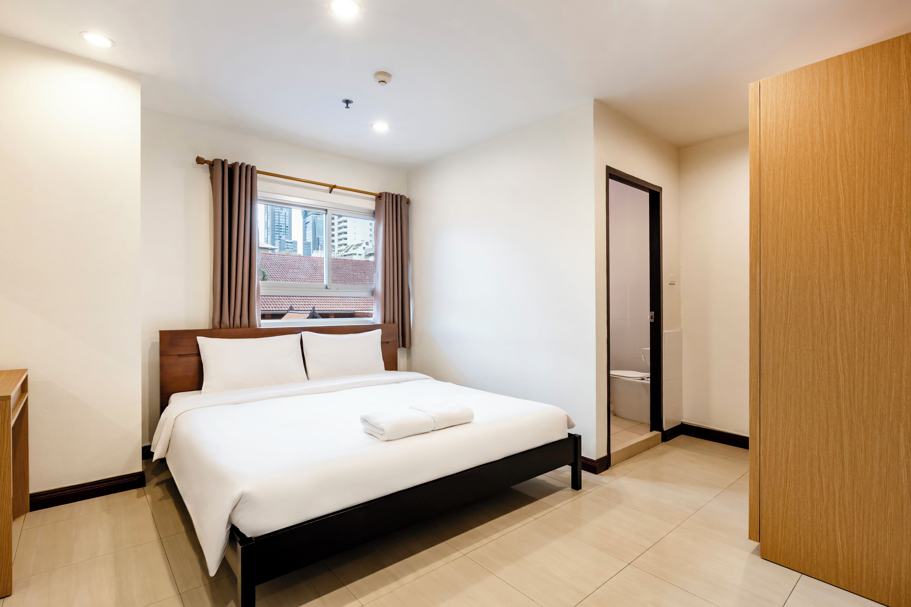 Une chambre d’hôtel avec un lit, une fenêtre avec des rideaux, une armoire en bois et une porte donnant sur une salle de bain.