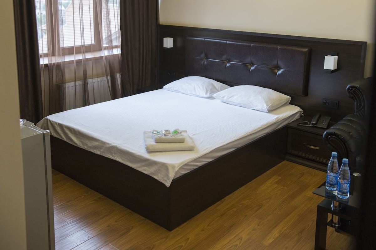 Una cama bien hecha en una habitación de hotel, con piso de madera, una mesita de noche con lámpara y una ventana con cortinas.