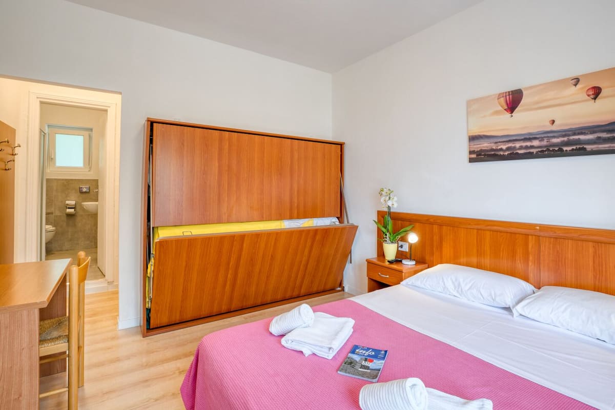 A hotel room with a bed, a wooden wardrobe, a desk, and a bathroom visible through an open door.