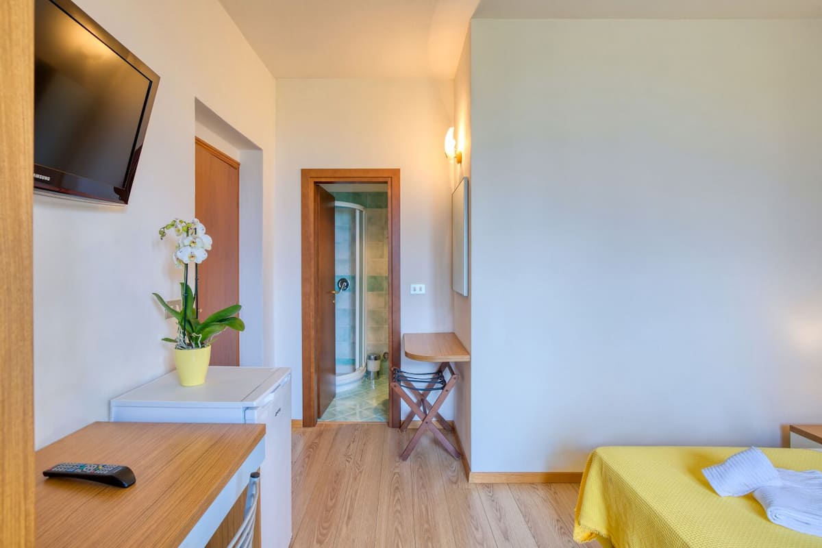 A hotel room with a wooden desk, a TV mounted on the wall, a bed with a yellow bedspread, and a shower area visible through an open door.