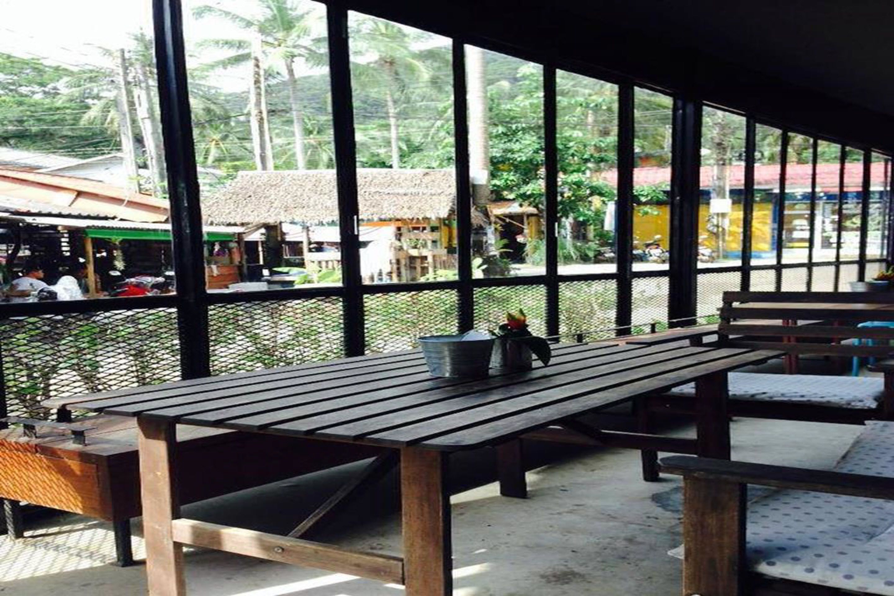 An outdoor seating area with wooden benches and tables, large windows offering views of a village with thatched-roof buildings and palm trees.