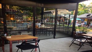 An outdoor seating area of a coffee shop with tables and chairs, a glass wall with a sign, and a view of the surrounding area. - Sleepy Owl Hostel (Ko Chang)