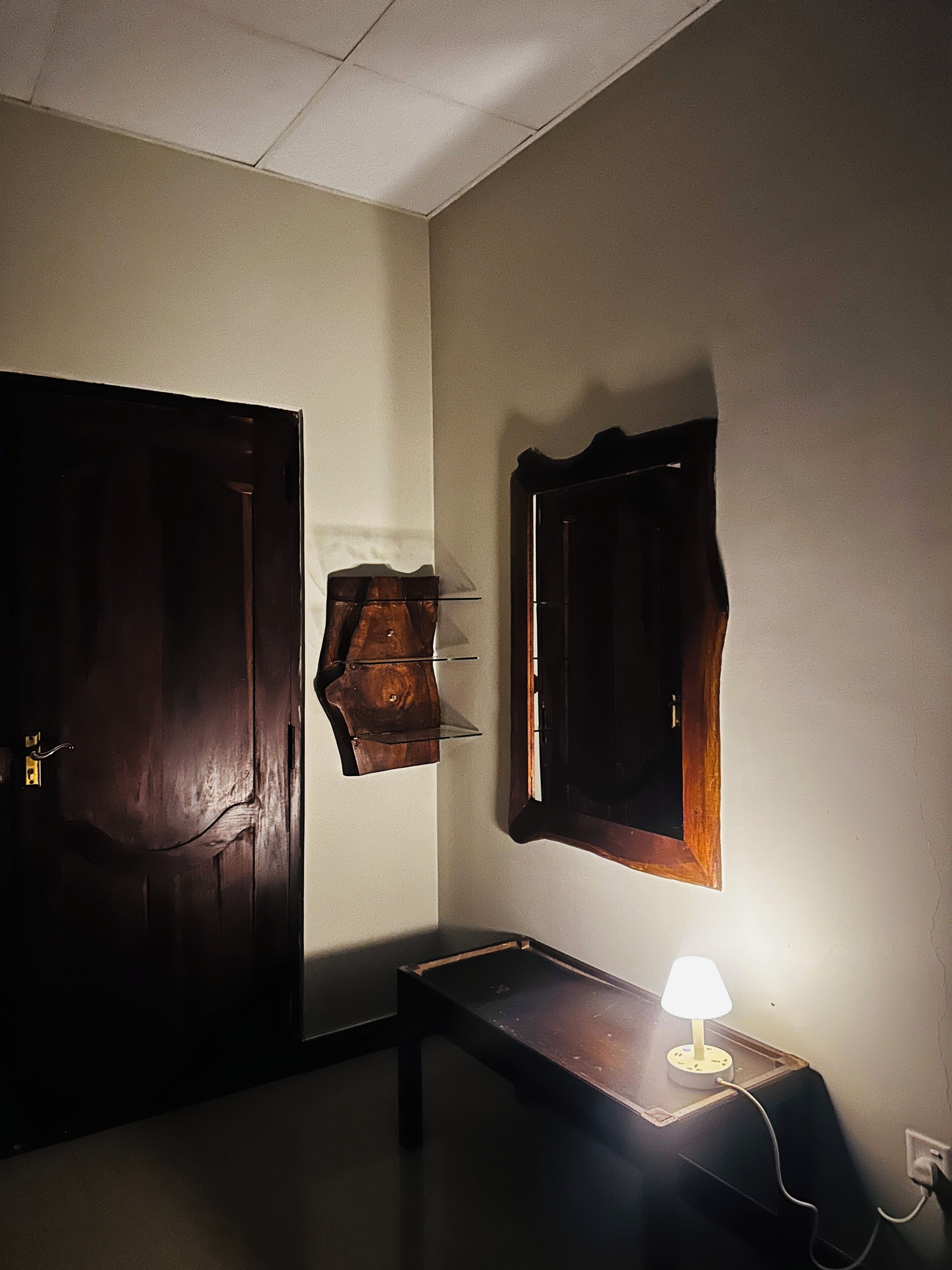 A room with a wooden door, a wall-mounted mirror, a small table with a lamp, and a wall socket.