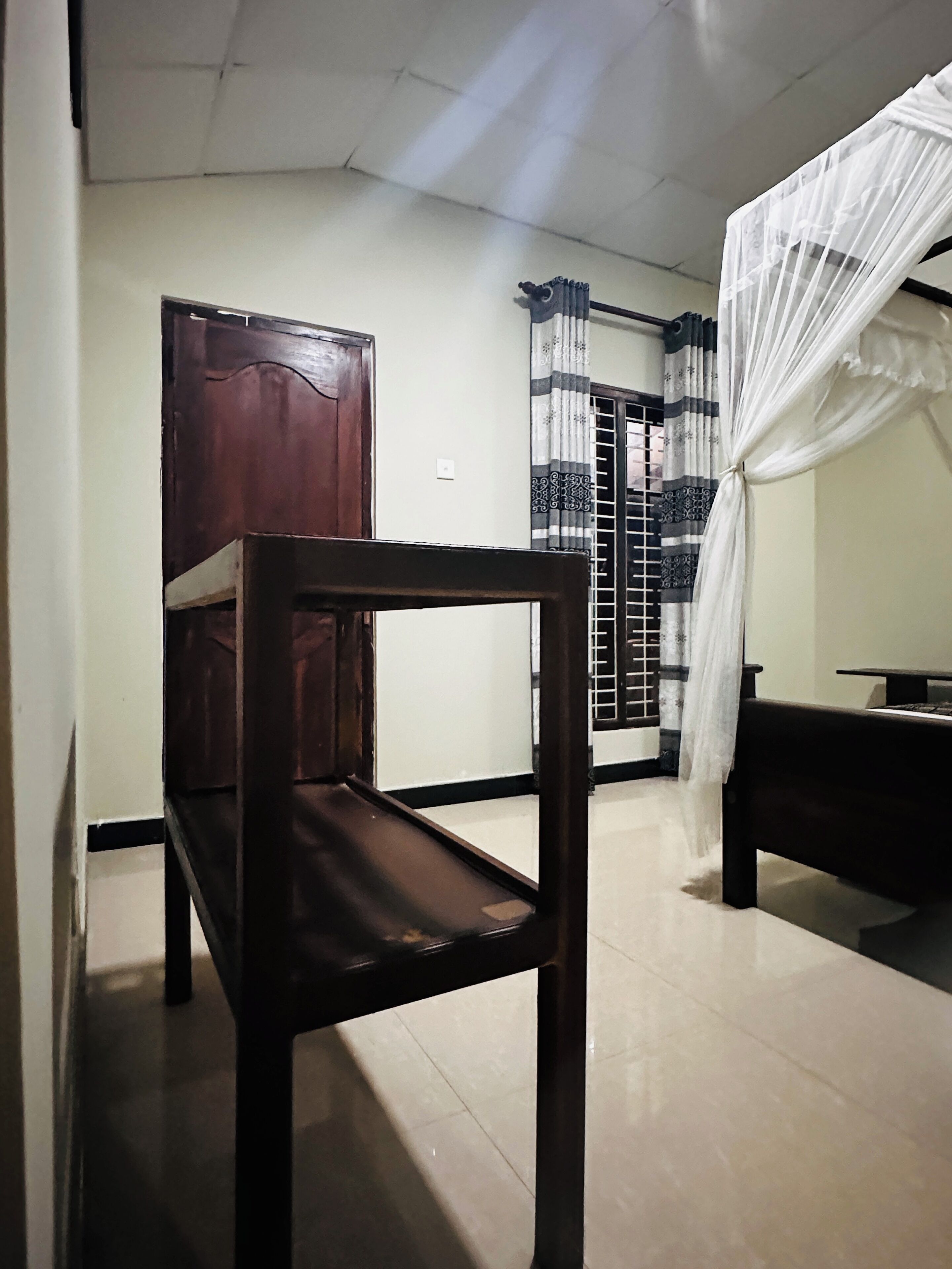 A room with a wooden chair, a bed with a white canopy, a window with patterned curtains, and a door.
