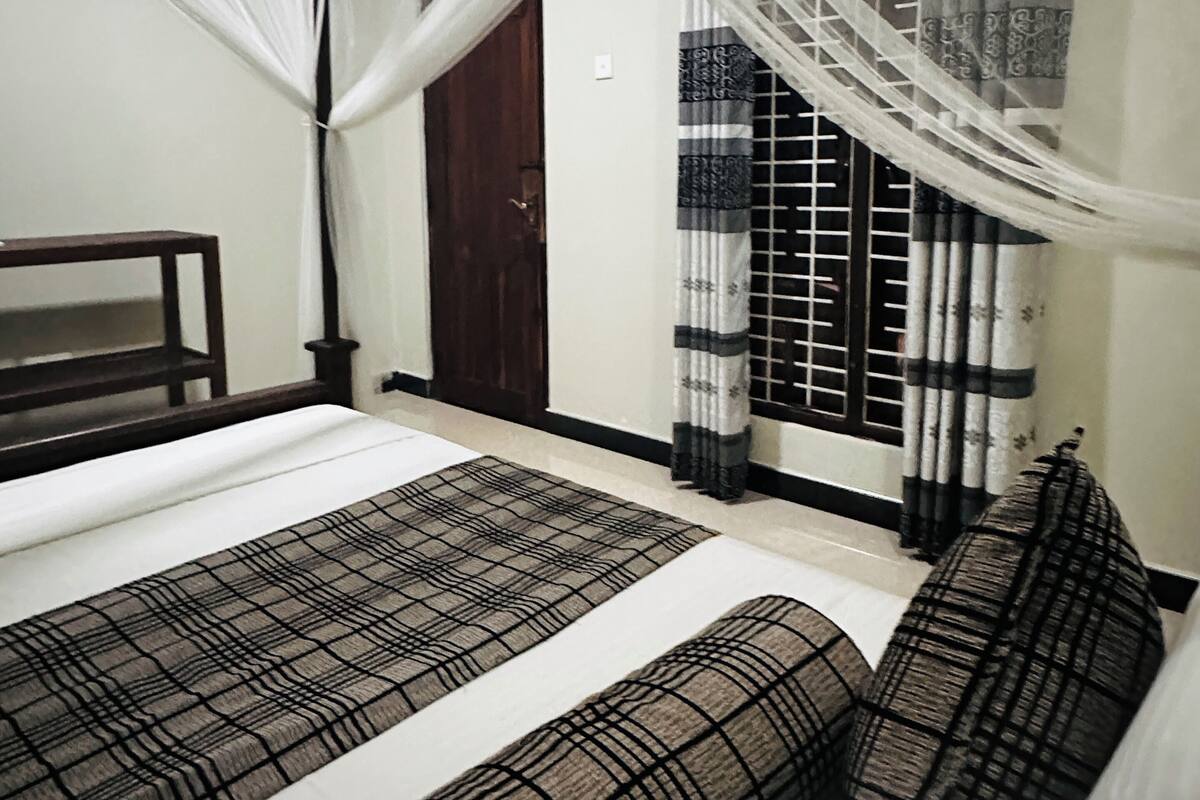 Um quarto com uma cama, um banco de madeira, uma porta e uma janela com cortinas.