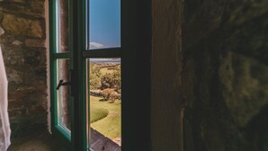Vista a través d'una finestra amb marc verd, que mostra un mur de pedra i un paisatge amb arbres i cel blau.