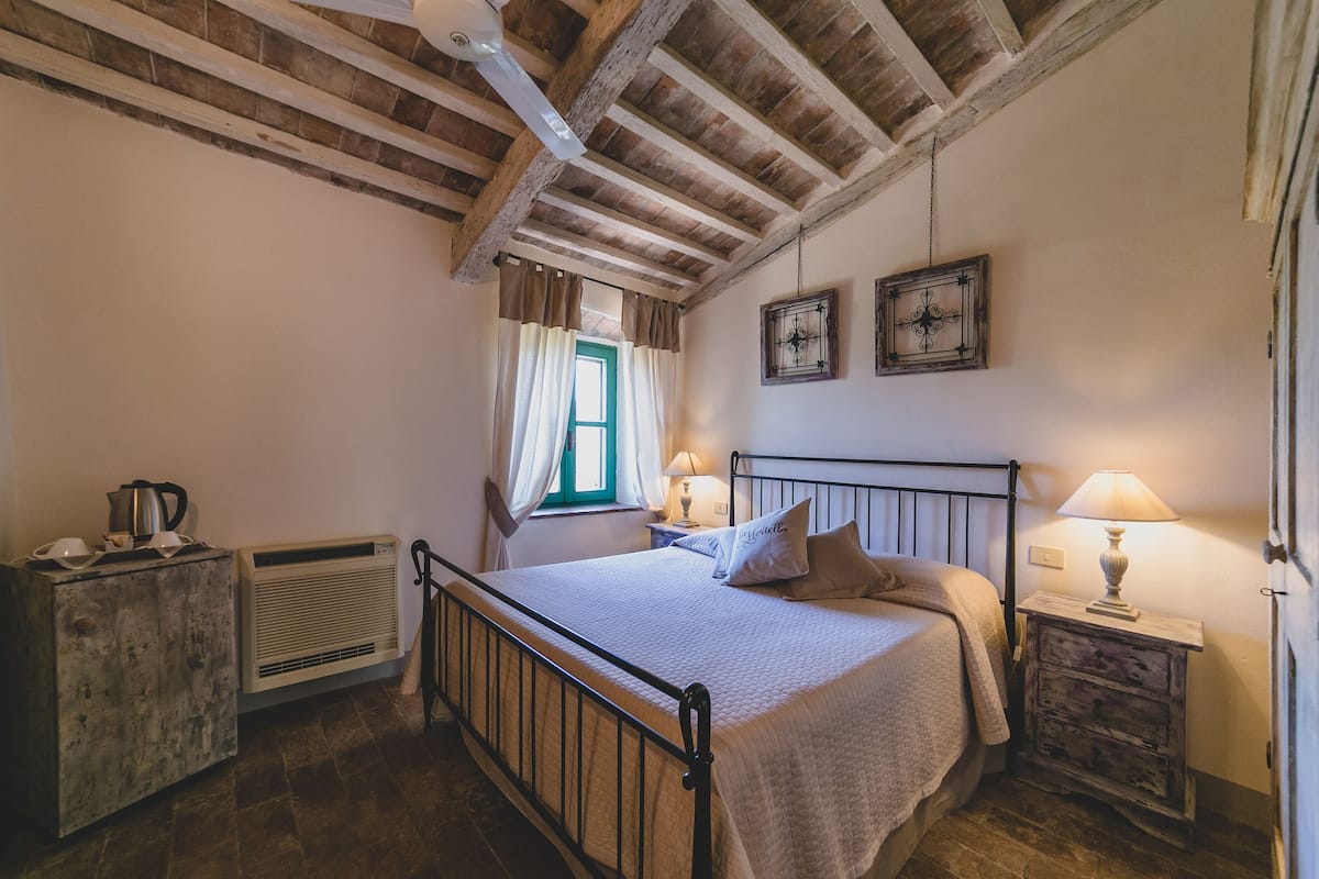 Una camera da letto con un letto, comodini, una finestra con tende, un termosifone e una ventola a soffitto.