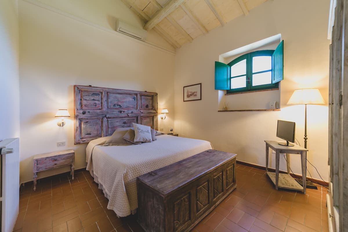 Una camera da letto con una testiera in legno, un letto con biancheria da letto bianca, un comodino, una lampada da letto, un tavolino con una lampada e una finestra con persiane blu.