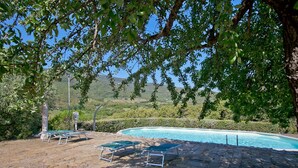 Una piscina amb gandules i una zona pavimentada, envoltada de vegetació i turons al fons.