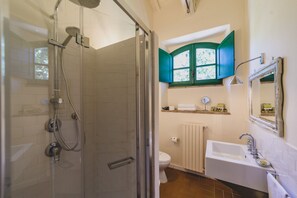 A bathroom with a glass-enclosed shower, a sink, a toilet, and a window with green shutters. - Country Relais la Mortella (Monte Argentario)