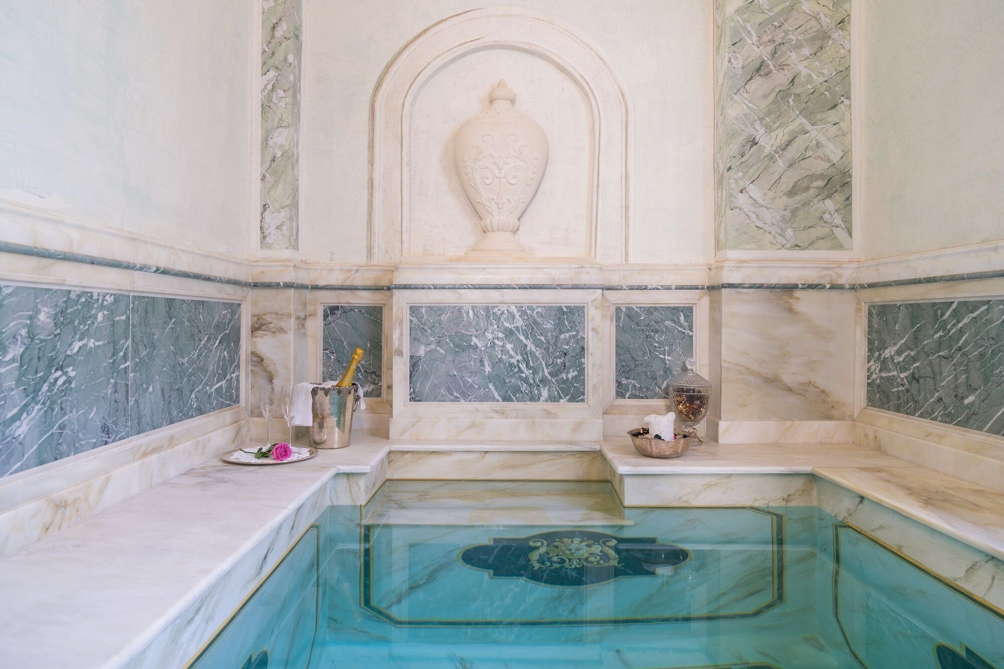 Un baño con suelo de mármol, una bañera integrada, un jarrón decorativo y una botella de champán.