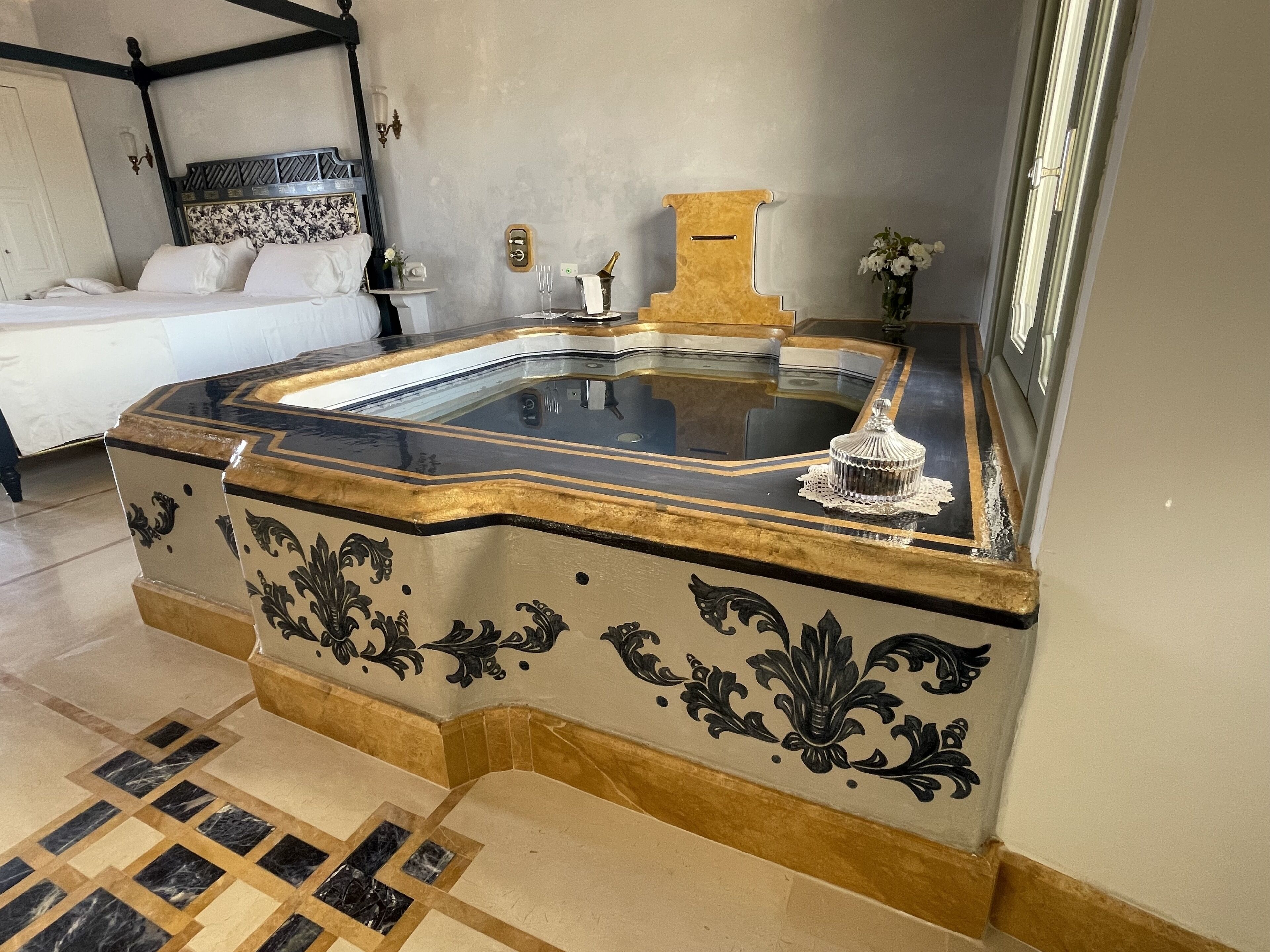 Ein Schlafzimmer mit einem großen Bett, einem Nachttisch, einem Zier-Spiegel und einem kleinen Tisch mit Blumen in einer Vase.