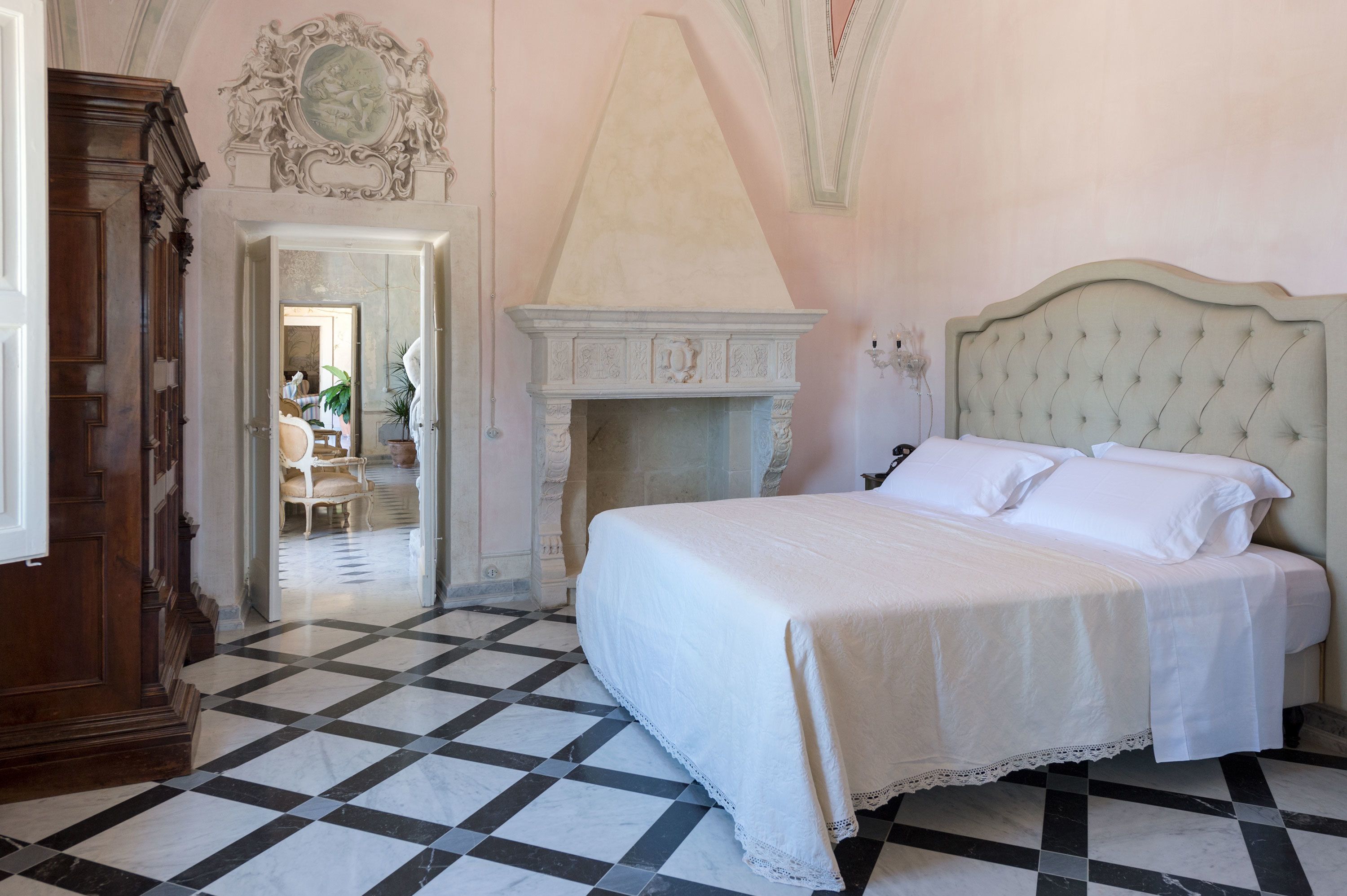 A bedroom with a checkered floor, a bed with white linens, a fireplace, and a mirror above the bed.