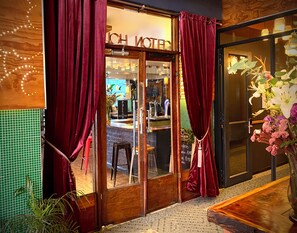 Entrada de un hotel con puerta de madera, cortinas rojas, un letrero que dice "HOTEL" y suelo de baldosas.