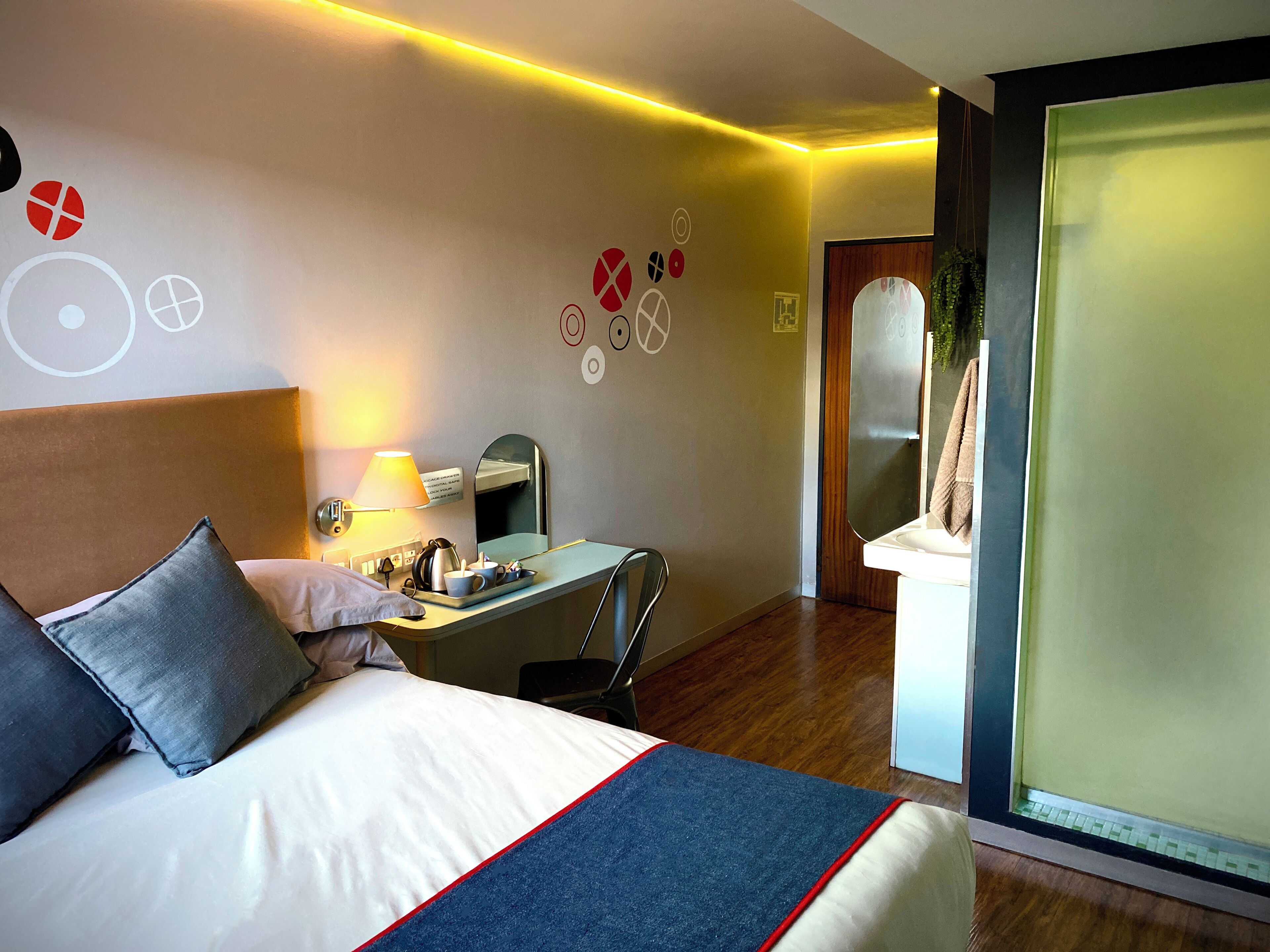 A hotel room with a bed, a desk, a lamp, a mirror, and a bathroom visible through a frosted glass door.