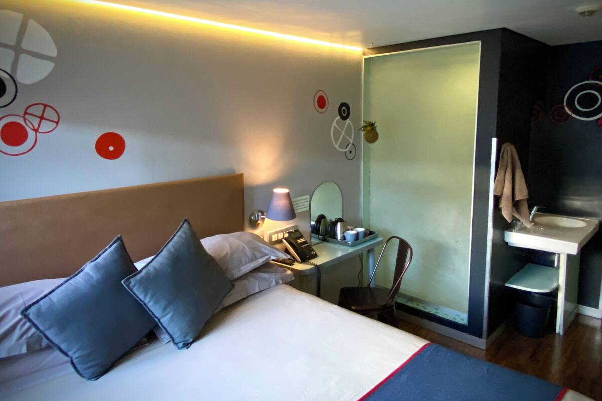 Une chambre d’hôtel comprenant un lit, une table de chevet, une lampe, un miroir et une petite salle de bain avec un lavabo et un miroir.