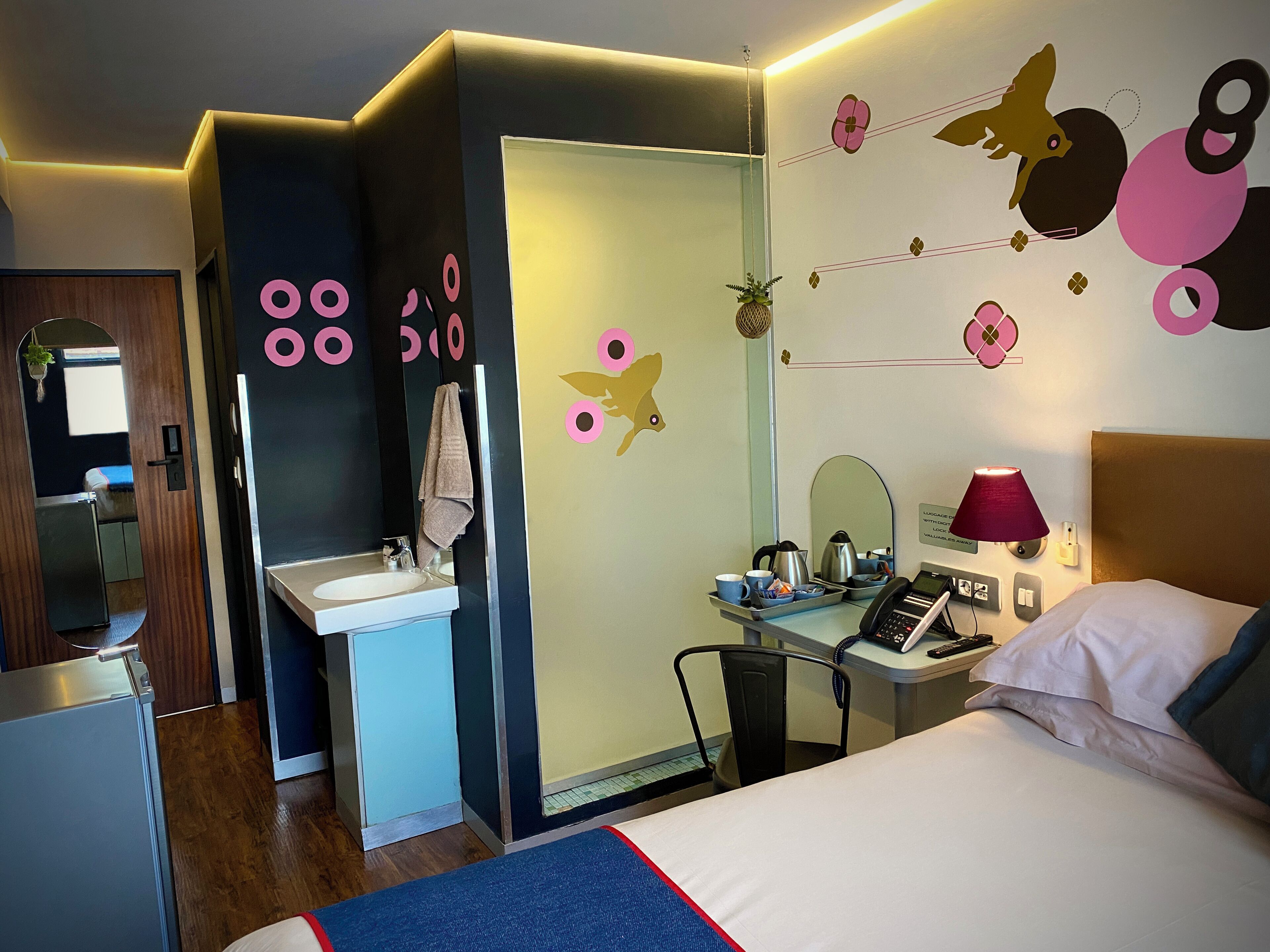 A hotel room with a bed, a desk with a lamp, a chair, a desk lamp, a phone, and a small table with a kettle and cups.