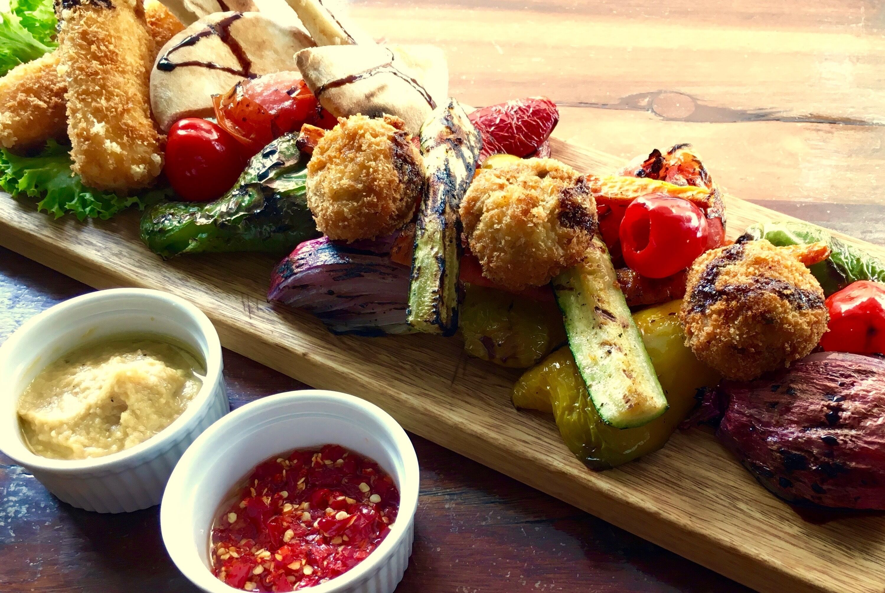 Una tabla de madera con verduras a la parrilla, carne y productos empanizados, acompañada de dos salsas para mojar.