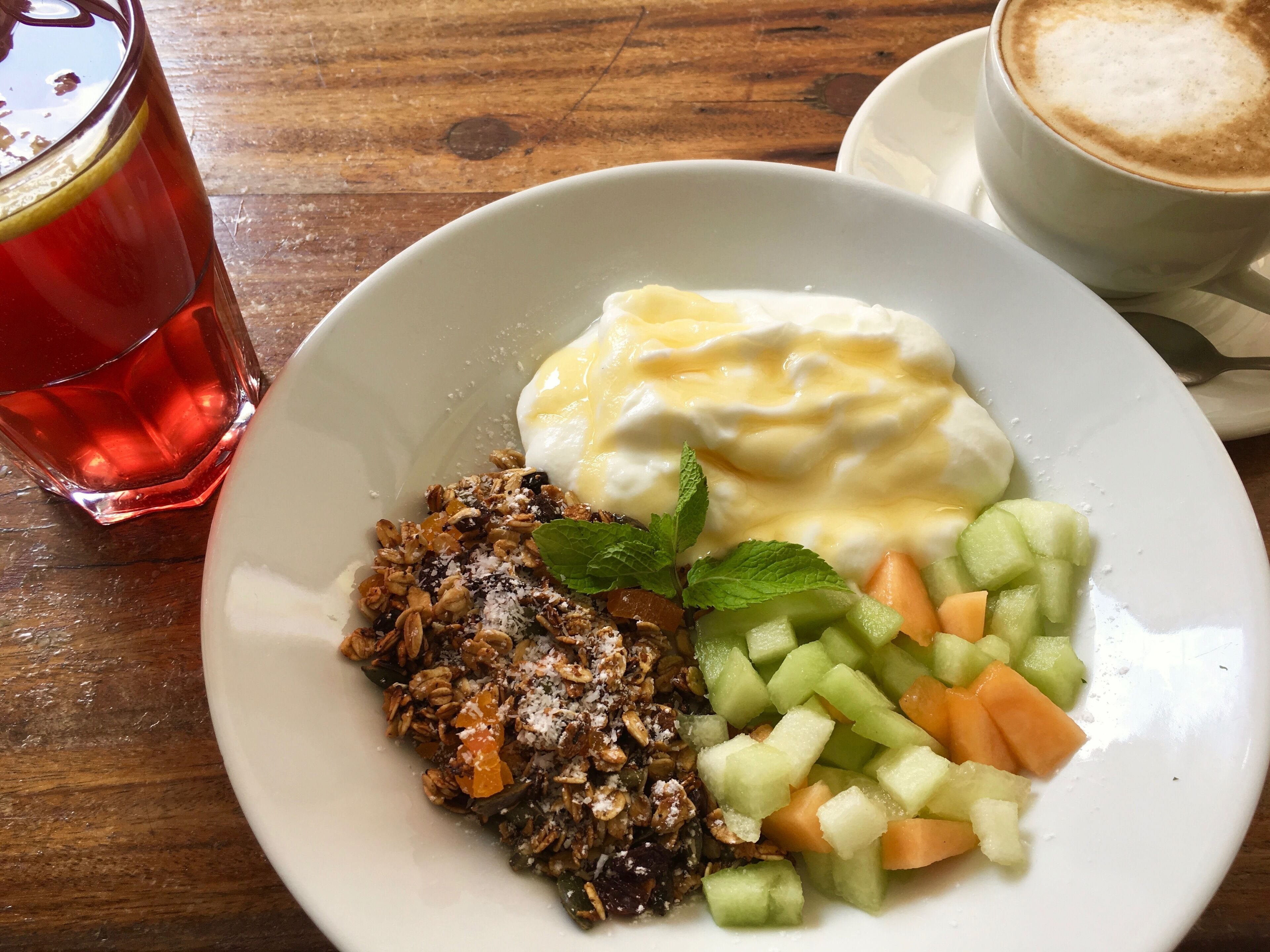 A plate with yogurt, granola, and diced fruits, a glass of red beverage, and a cup of coffee on a wooden table.