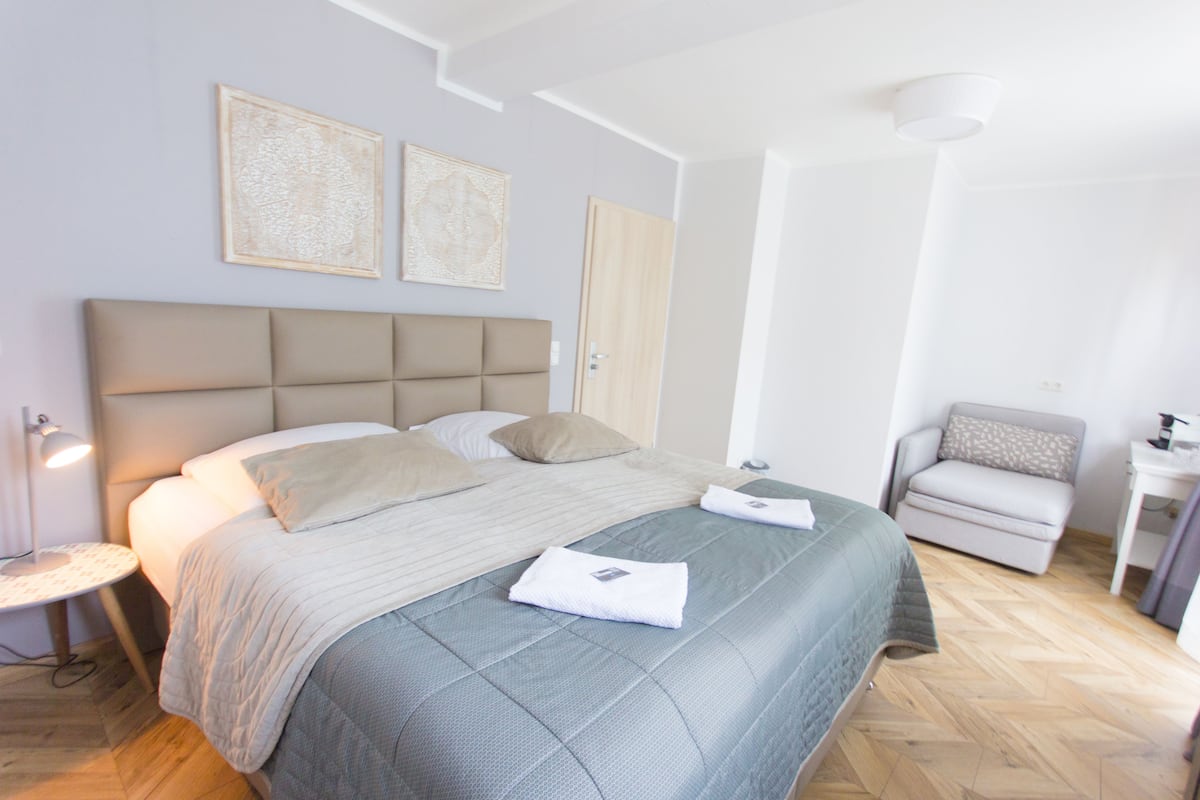 A neatly arranged bedroom with a large bed, a bedside table, a chair, and two framed artworks on the wall.