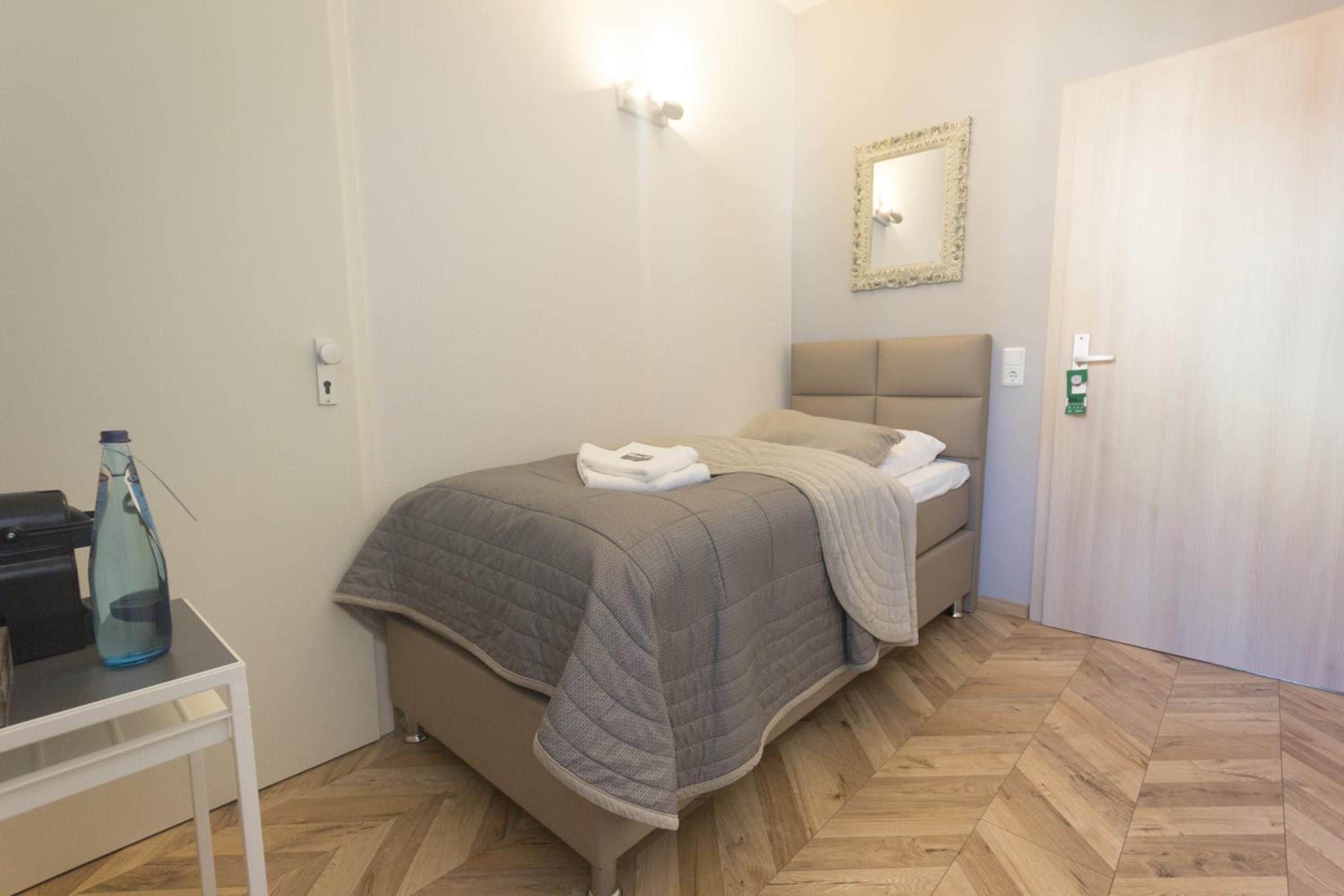A bedroom with a bed, a bedside table with a bottle, a mirror on the wall, and a door.