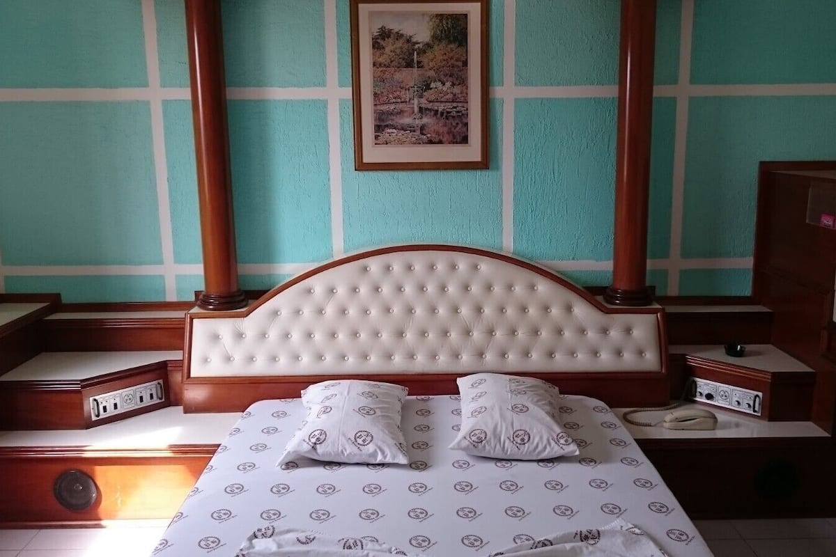 Une chambre à coucher avec une tête de lit capitonnée, deux tables de chevet, un lit avec une literie à motifs et un tableau encadré au mur.