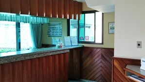 Un espace d’accueil d’hôtel comprenant un comptoir, une fenêtre avec des rideaux, un panneau d’affichage indiquant les tarifs des chambres et un panneau mural en bois.