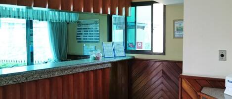 A hotel reception area with a counter, a window with curtains, a bulletin board with room rates, and a wooden wall panel.