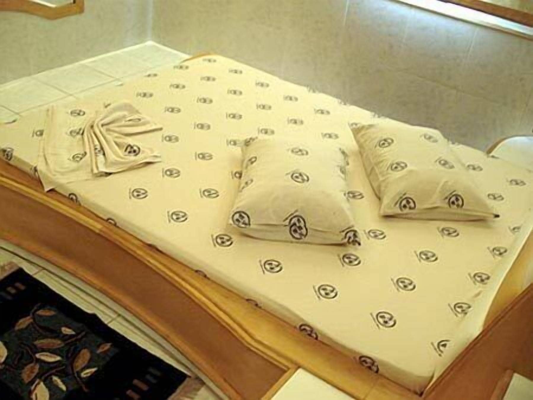 Cama com colcha estampada e almofadas, cabeceira de madeira e tapete escuro com padrão.