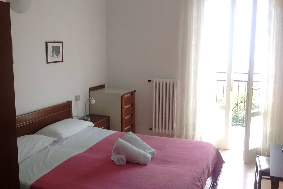 Un dormitori amb llit, una tauleta de nit, un prestatge, un radiador i una finestra amb cortines.