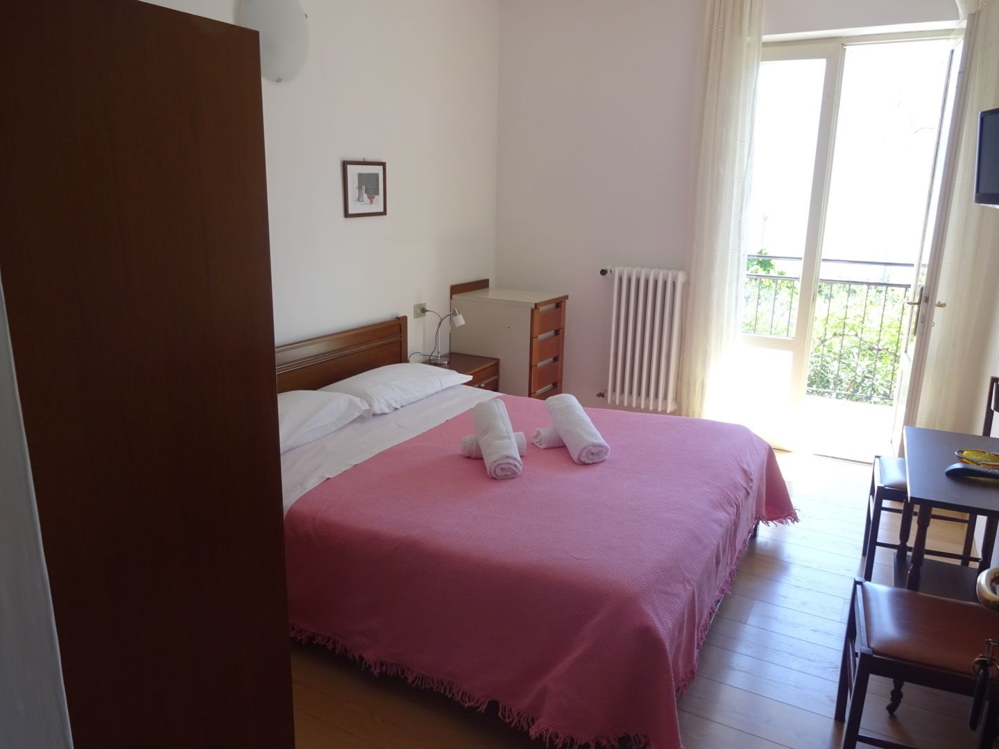 Habitación de hotel con cama, mesita de noche, lámpara, radiador y vistas al exterior.