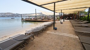 A beachside terrace with sun loungers and picnic tables, overlooking a calm sea and a distant town. - Casamia Boutique Hotel (Bodrum)