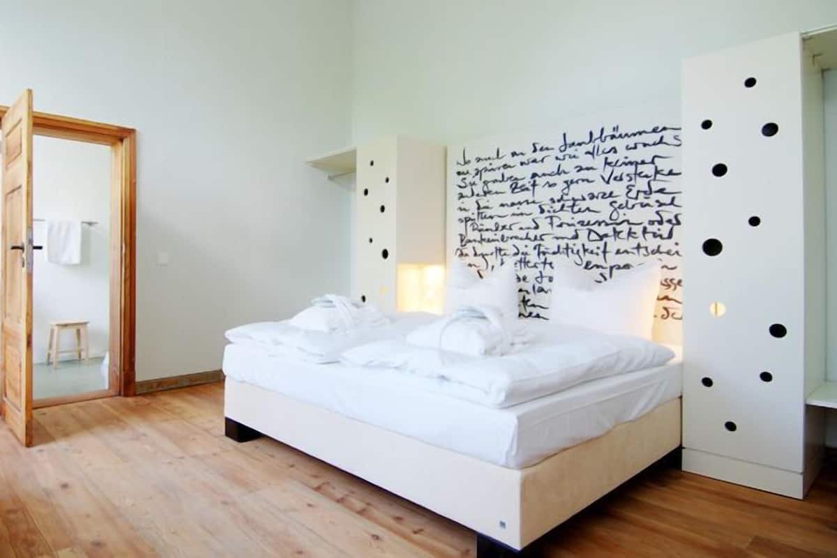 Une chambre avec un plancher en bois, un lit recouvert de draps blancs, une porte en bois et un mur orné d’ouvertures circulaires noires.
