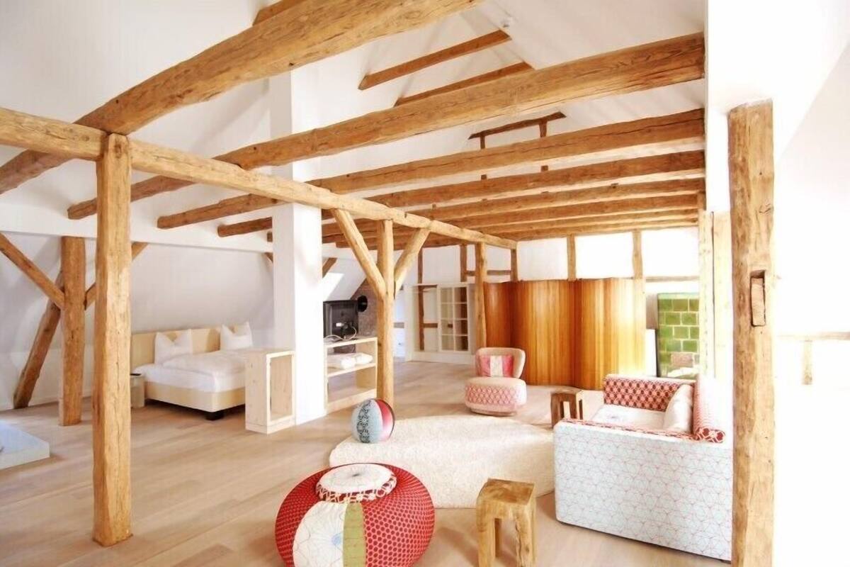 Une chambre spacieuse aménagée en mezzanine, avec des poutres apparentes, un lit, un coin salon avec des coussins et un petit tabouret en bois.