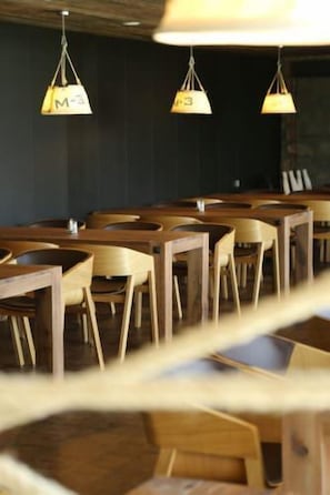 A restaurant interior with wooden tables and chairs, hanging pendant lights, and a dark wall in the background. - Alsóörs Marina Hotel (Alsoors)