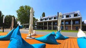A wooden deck with blue cushions and white umbrellas, a building with a sloped roof and solar panels, and a clear blue sky. - Alsóörs Marina Hotel (Alsoors)