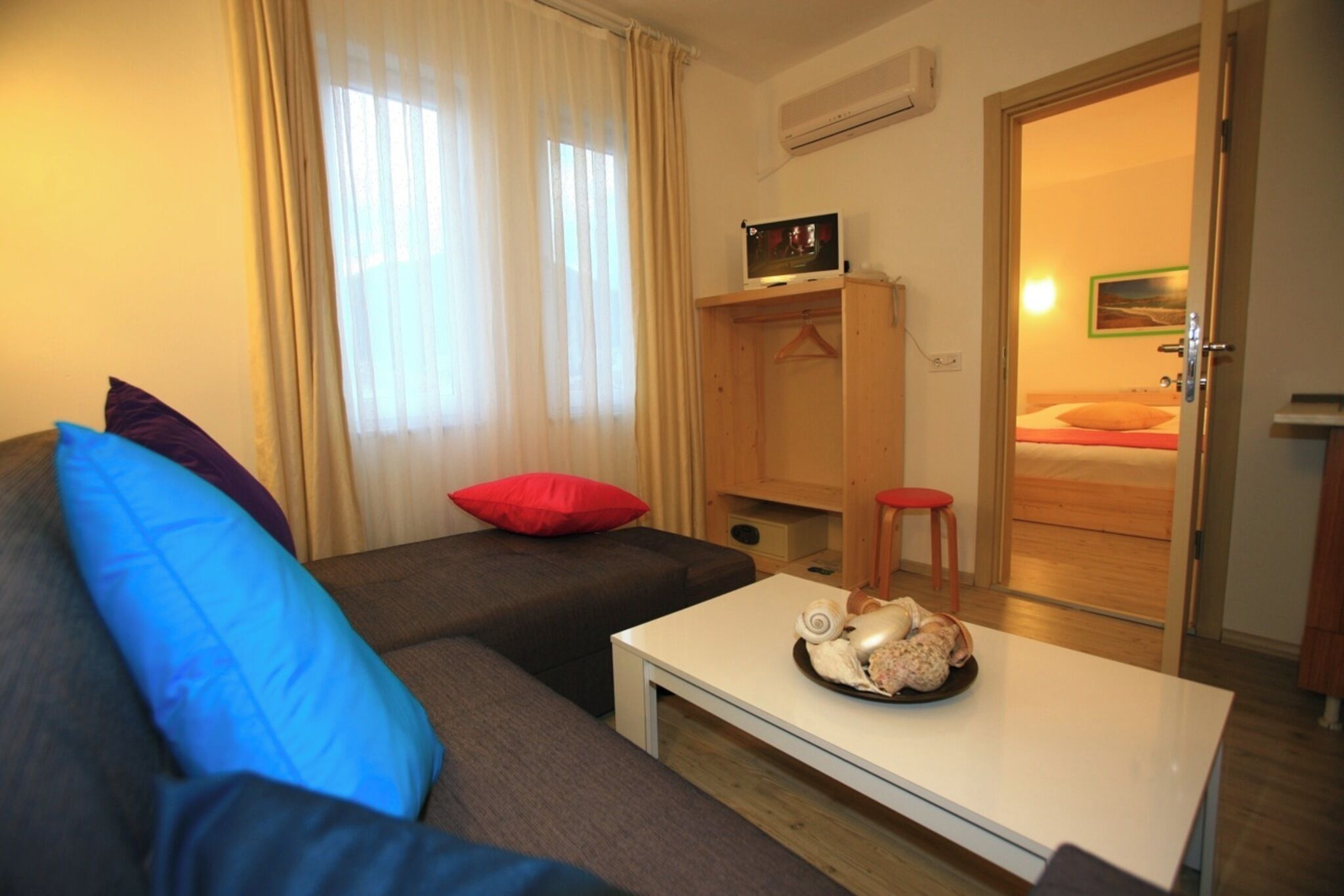 Une pièce comprenant un canapé, une petite table ornée d’un objet décoratif, une armoire en bois et une porte donnant sur une chambre.
