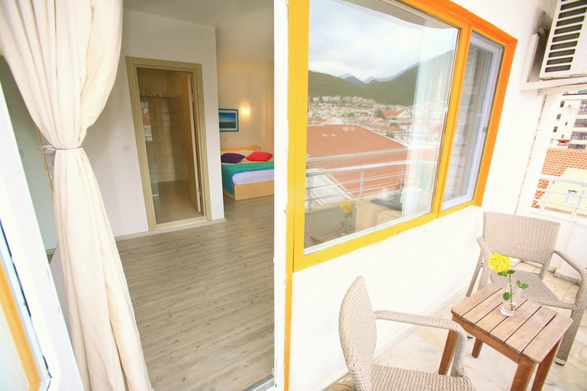 Un balcon donnant sur la ville et les montagnes, une table en bois avec un vase et une chaise en osier.