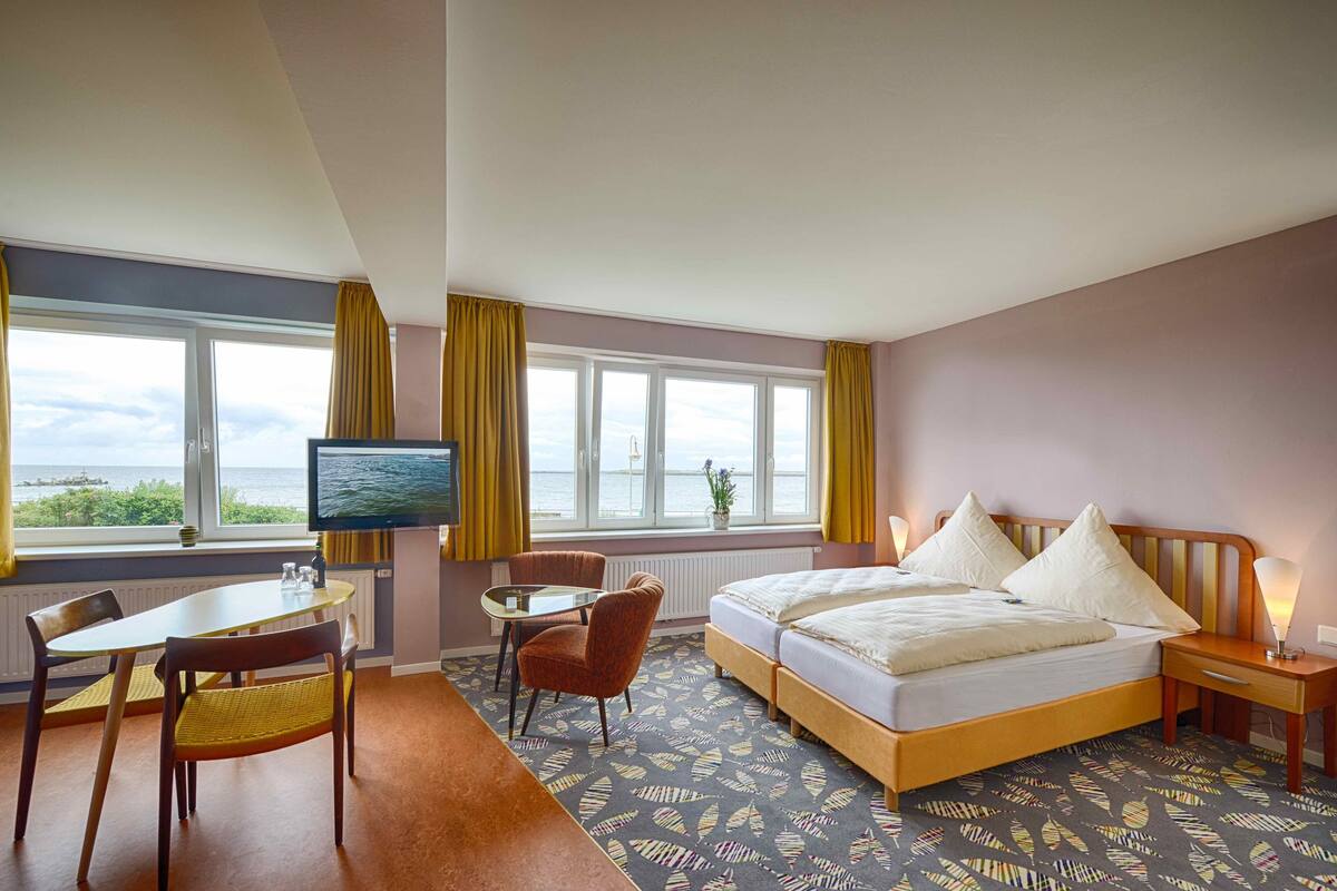 Habitación de hotel con cama, televisión, una mesa con sillas y vista al mar.