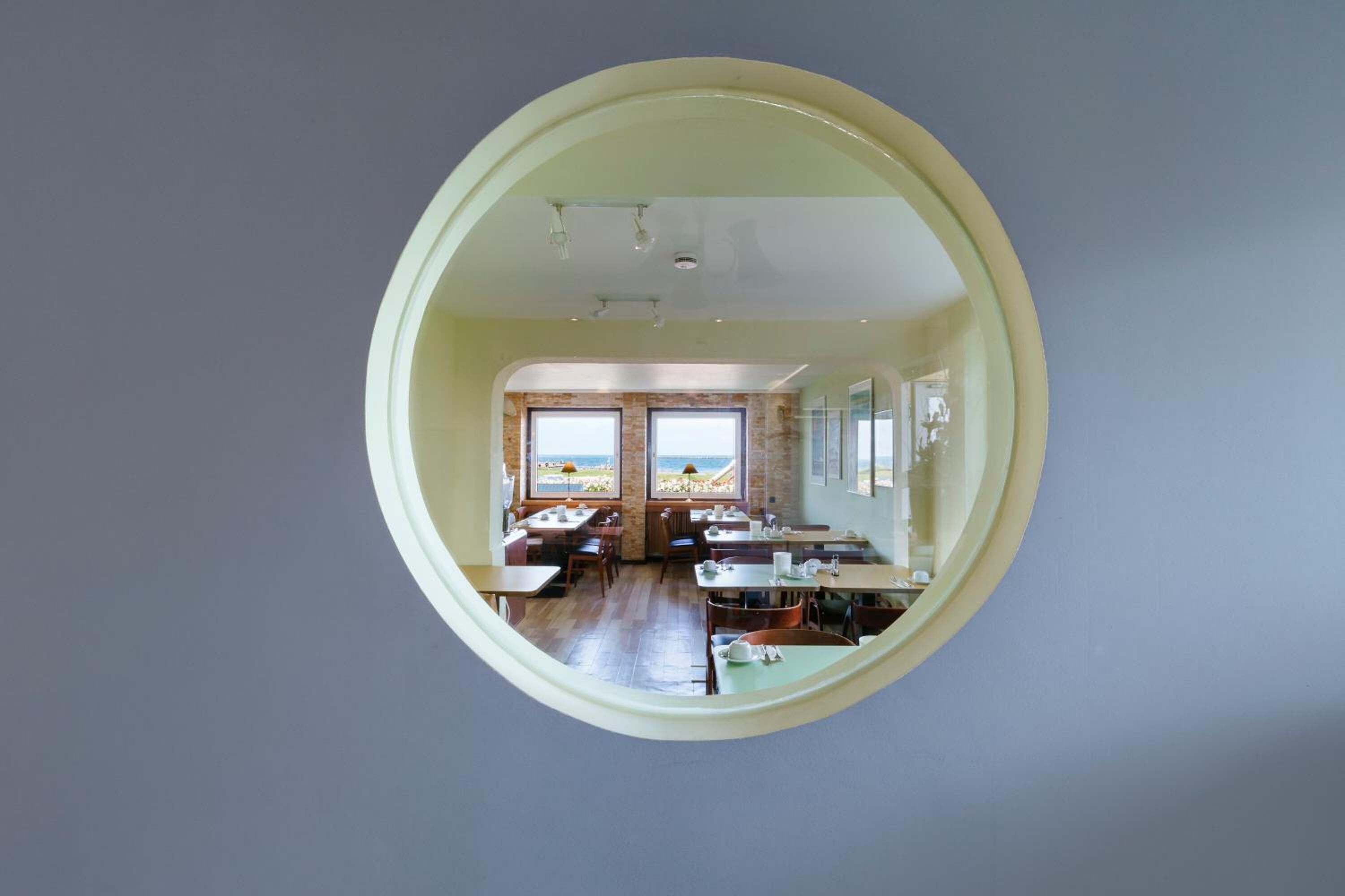 Un espace repas avec des tables et des chaises, de grandes fenêtres offrant une vue dégagée et un grand miroir rond accroché au mur.