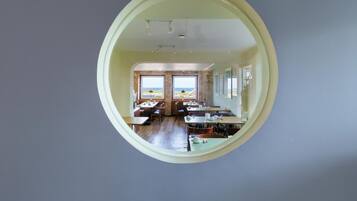 Un espace repas avec des tables et des chaises, de grandes fenêtres offrant une vue dégagée et un grand miroir rond accroché au mur.