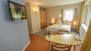Ein Hotelzimmer mit Bett, einem an der Wand montierten Fernseher, einem kleinen Tisch mit Fernbedienung und einem Fenster mit Vorhängen.