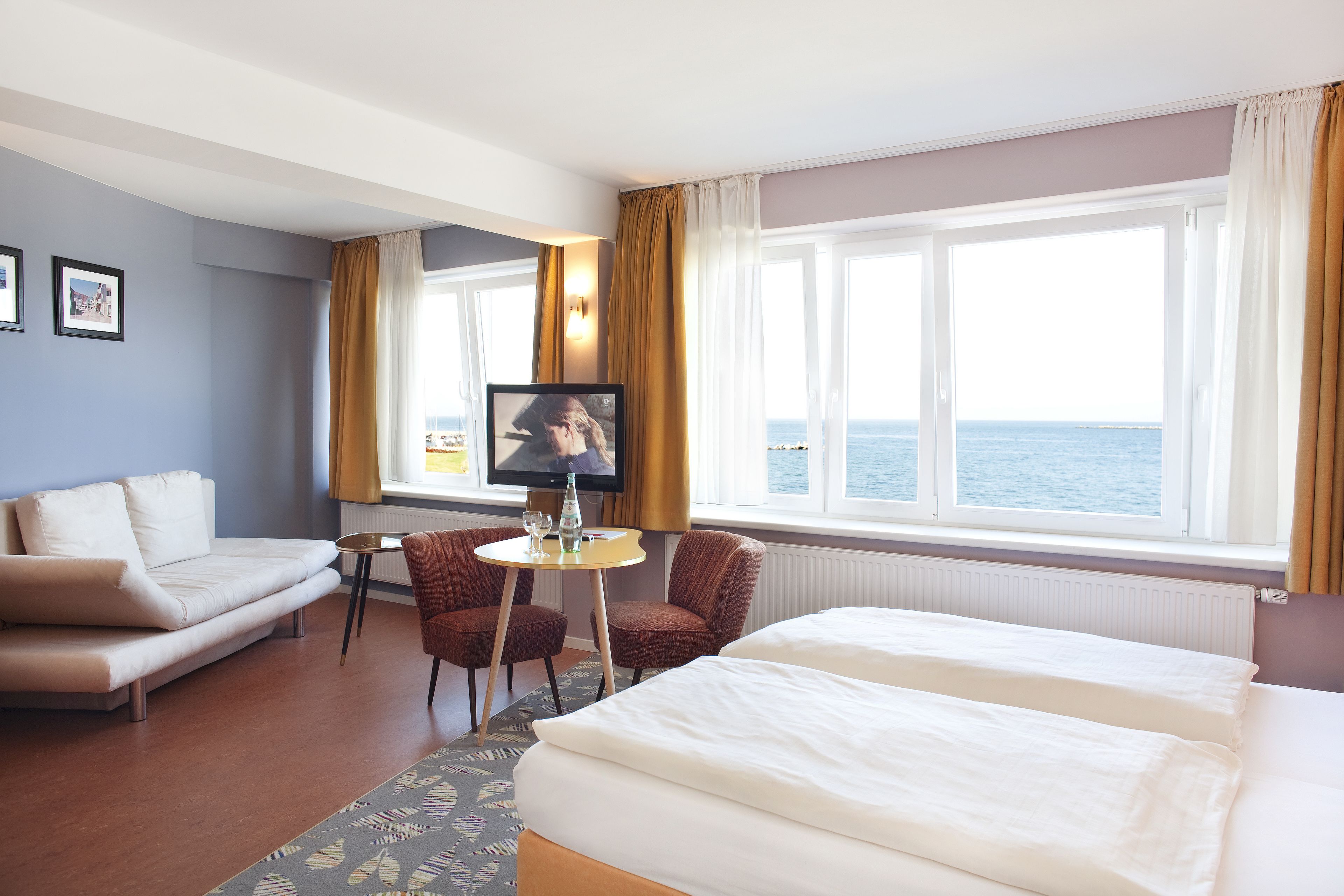 A hotel room with a bed, a sofa, a small table with a TV, and a view of the sea.