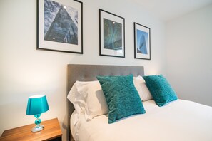 A neatly made bed with two teal pillows and a white pillow, a wooden nightstand with a teal lamp, and three framed pictures on the wall. - Bluestone Apartments - Didsbury (Manchester)
